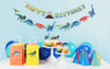 dinosaur birthday party table with dinosaur happy birthday banner, garland, cupcake toppers, gift bags, party hats and blue balloons